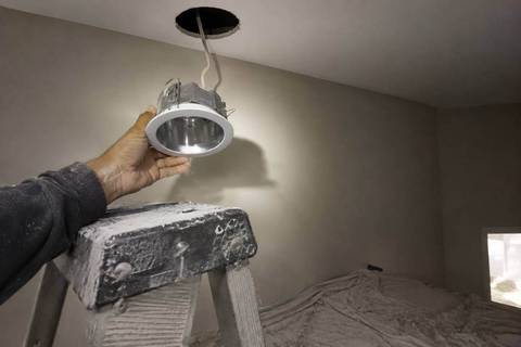 Circular opening cut in a high ceiling with a recessed housing held up to it, drywall dust on the ladder top
