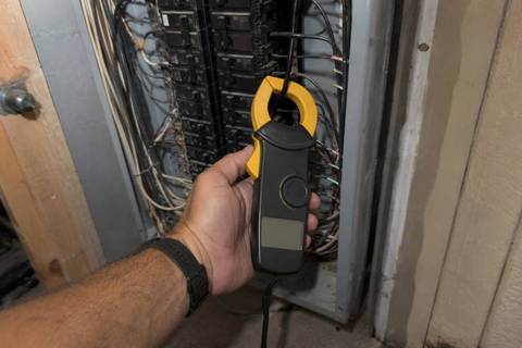 Clamp meter closed around a single insulated conductor inside an open panel, display angled toward the camera