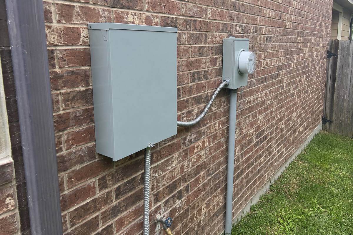 Gray transfer switch enclosure mounted on an exterior wall beside a meter, flexible conduit running between them